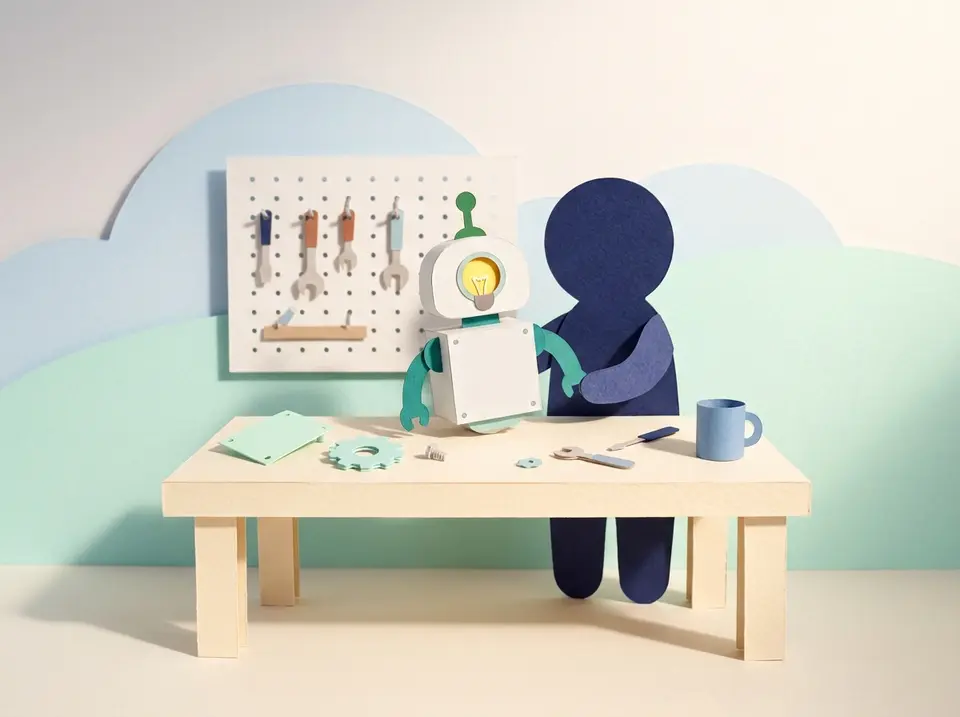 Paper-cutout diorama: a small paper figure stands at a workbench assembling a friendly little paper robot from parts, a pegboard of tools on the wall behind — the practical AI skills a team builds and keeps.